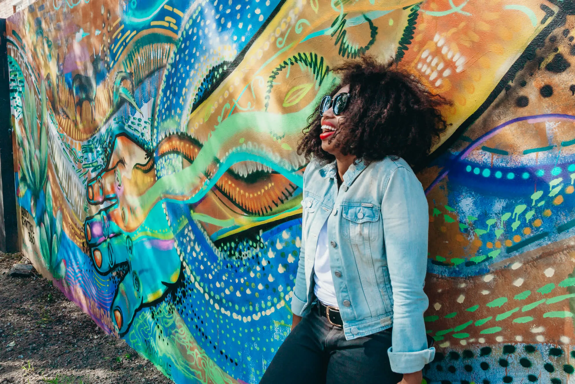 A woman in a denim jacket laughs in front of a vividly painted mural wall in Langa